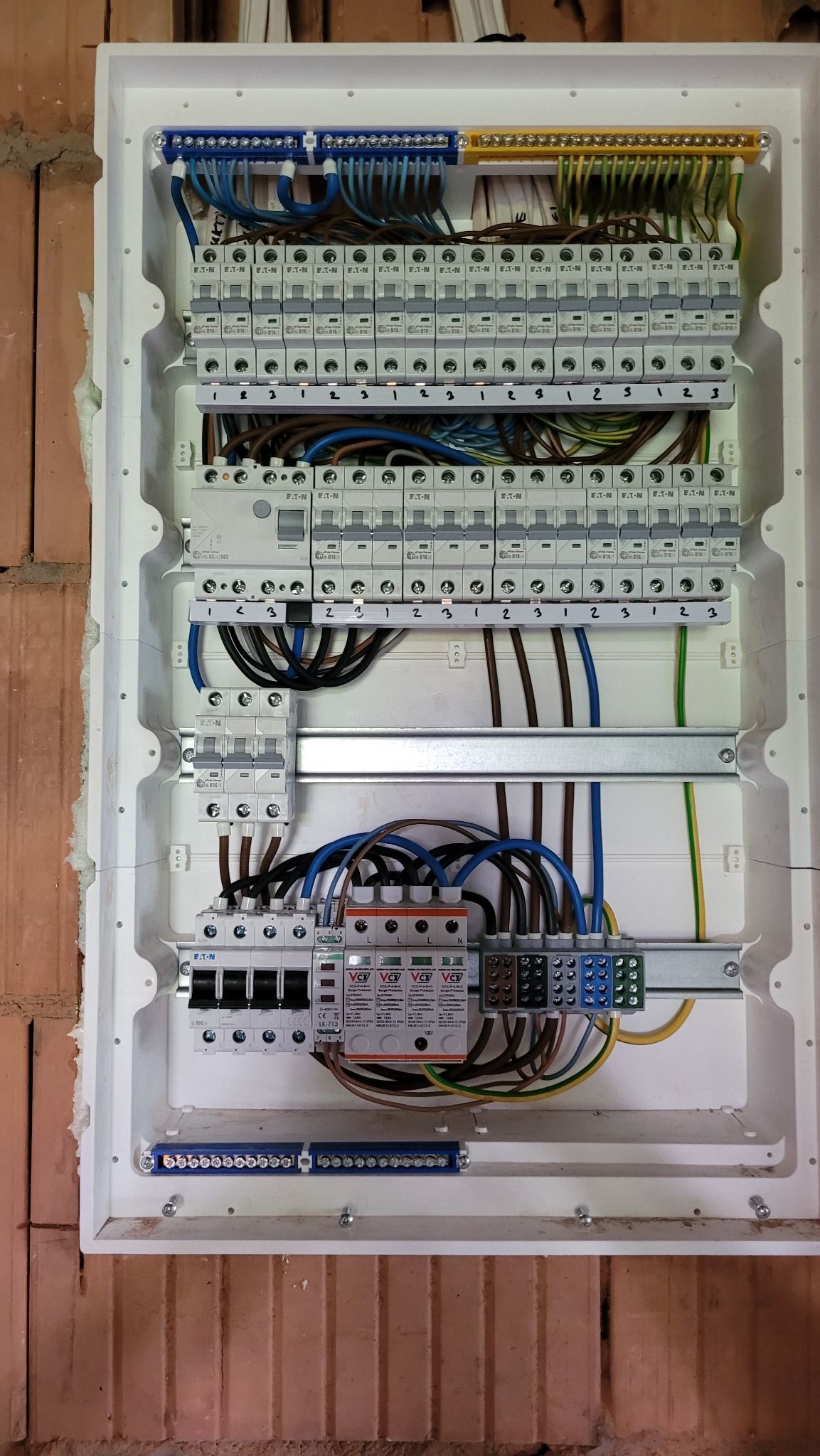 Otwarta skrzynka rozdzielcza z widocznymi zabezpieczeniami, przewodami o różnych kolorach i listwami zaciskowymi, zamontowana na ścianie z cegły.