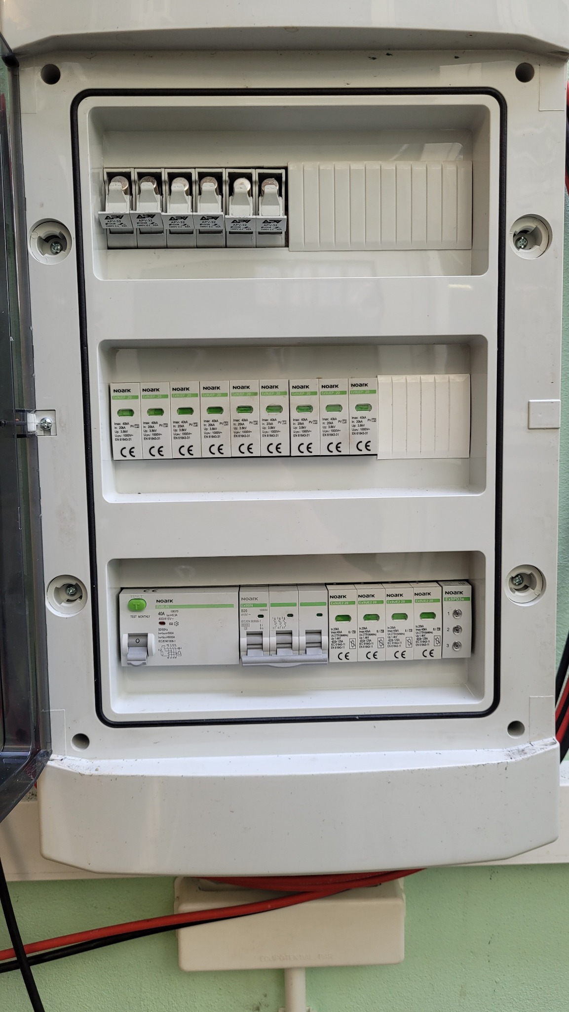 Otwarta skrzynka rozdzielcza z zamontowanymi bezpiecznikami i ogranicznikami przepięć marki Noark, widoczne przewody elektryczne.