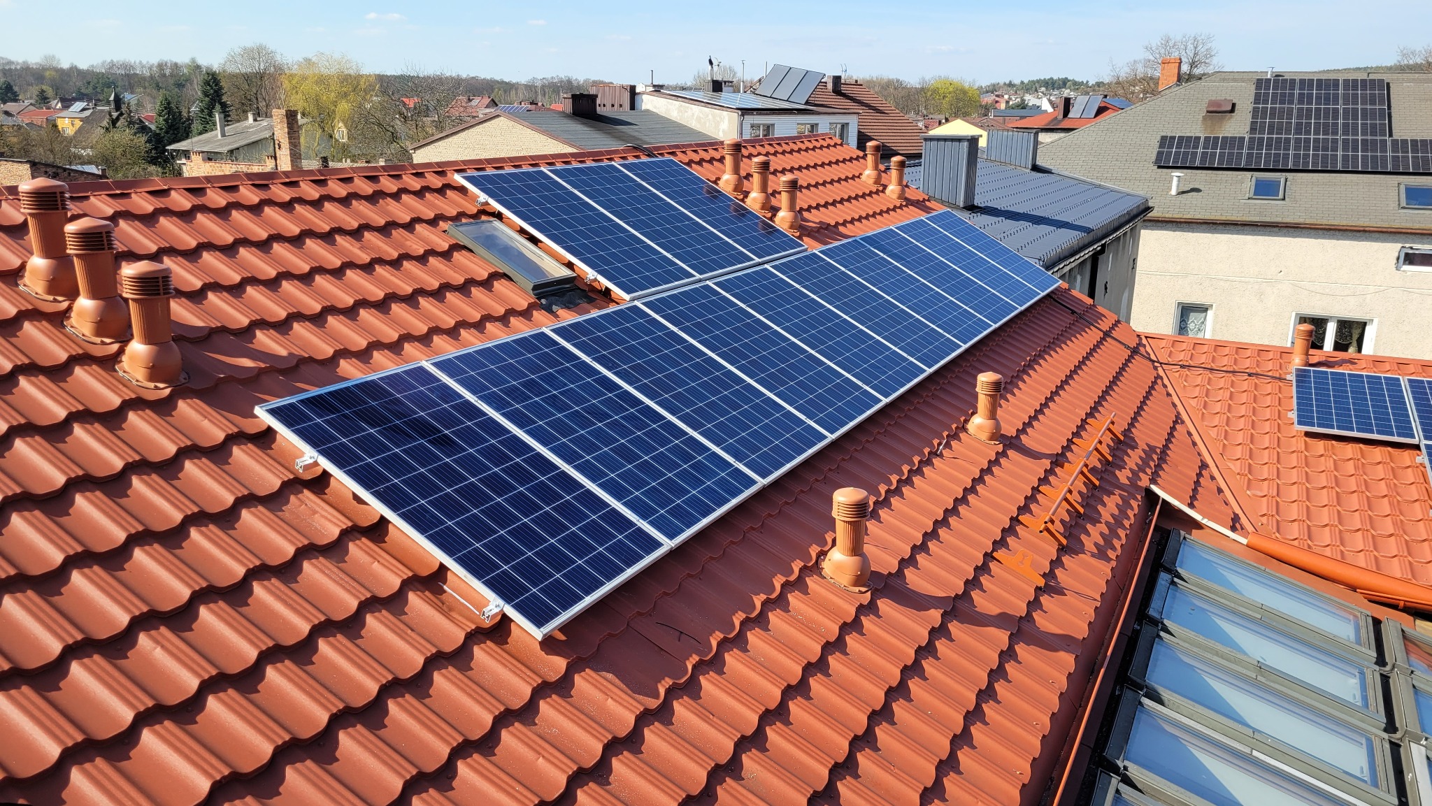 Panele fotowoltaiczne zamontowane na dachu z czerwonej dachówki ceramicznej z kominkami wentylacyjnymi; widoczne okna dachowe i fragmenty innych budynków w tle.