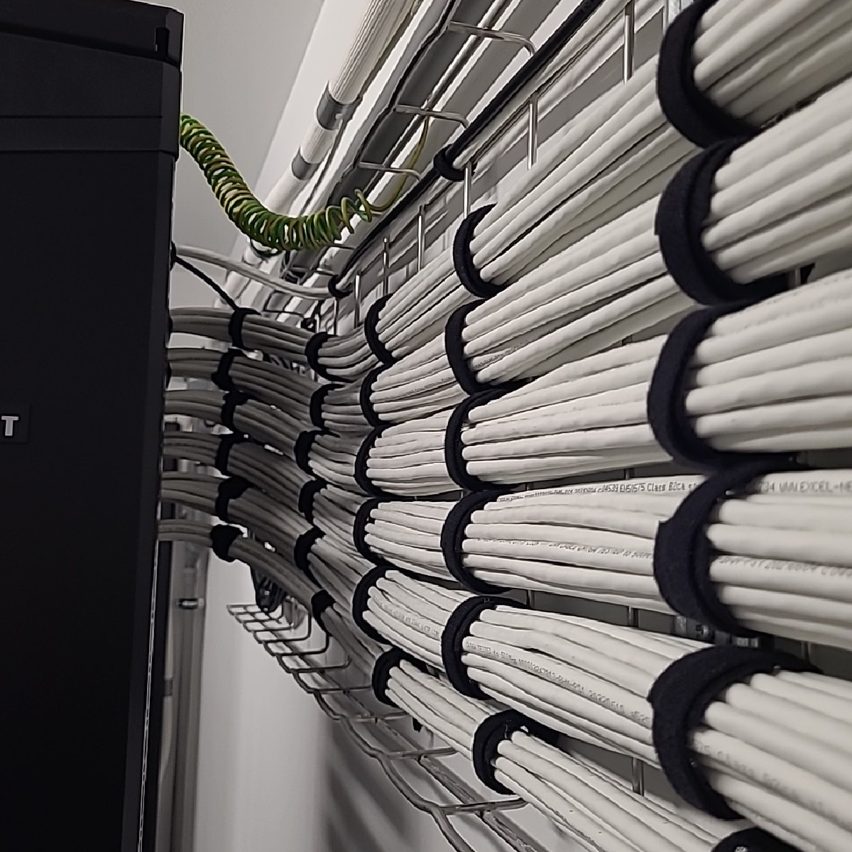 Równolegle ułożone kable w białej izolacji, zabezpieczone czarnymi opaskami i prowadzone na metalowej siatce, z widocznym kablem uziemiającym w kolorze zielono-żółtym, w tle ściana pomalowana...