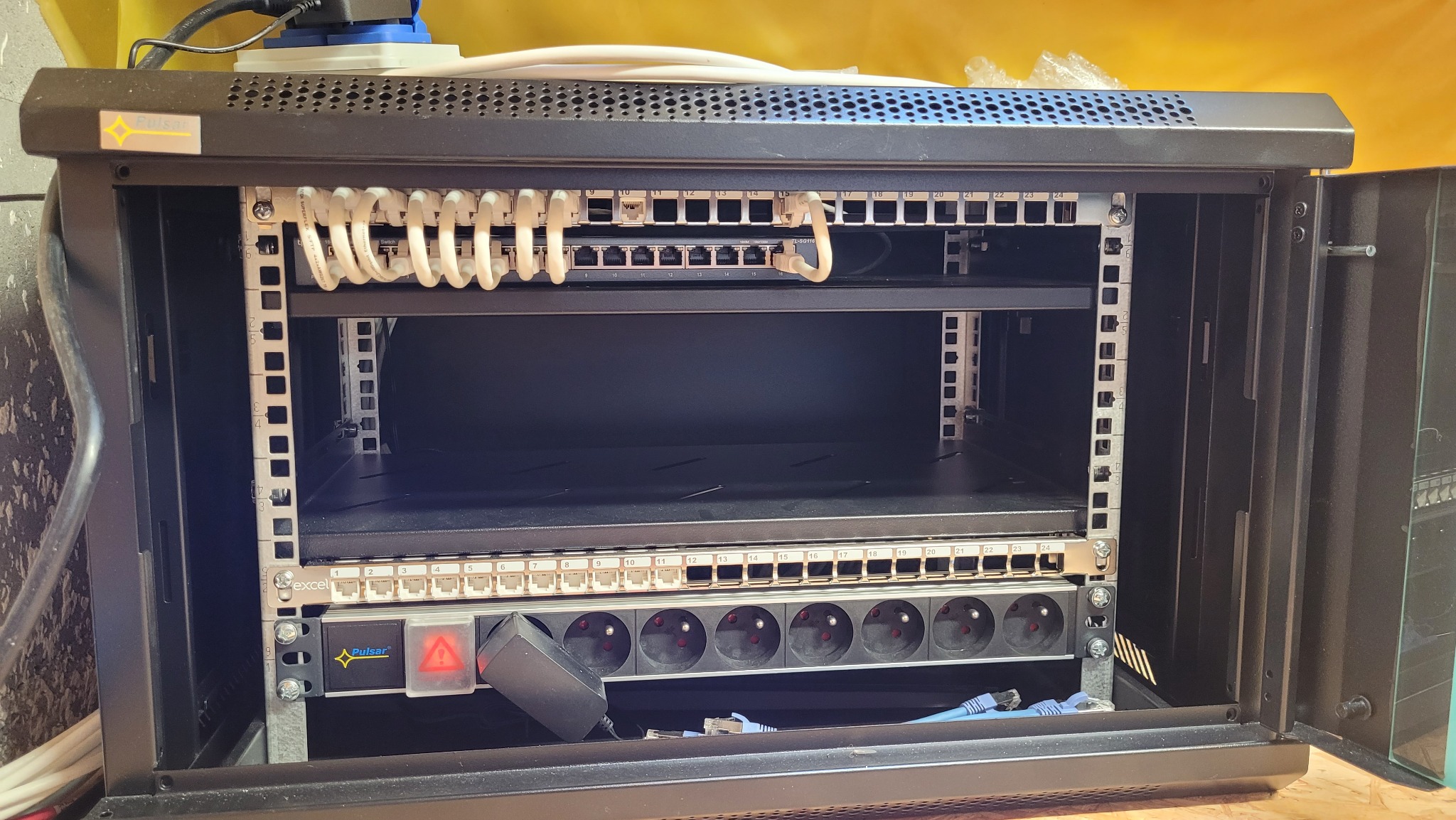 Otwarta szafa rack z panelami krosowniczymi, listwą zasilającą i uporządkowanymi kablami patchcord, widoczne oznaczenia portów i logo producenta Pulsar.