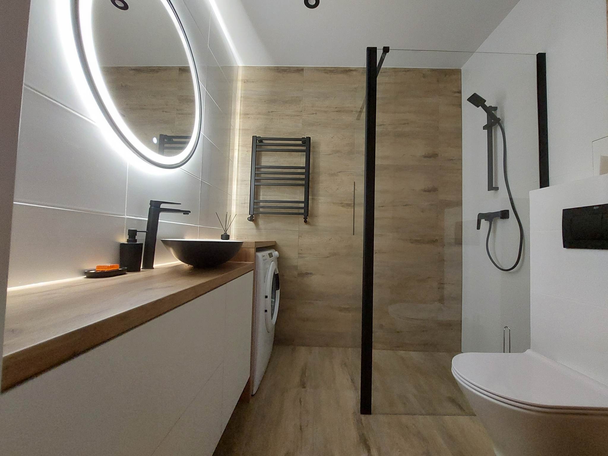 Nowoczesna łazienka z kabiną prysznicową typu walk-in, okrągłym lustrem z podświetleniem LED, czarną armaturą, drewnianym blatem i pralką w zabudowie.