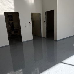 Piotr Cichoń - Szara, błyszcząca podłoga z żywicy w jasnym pomieszczeniu z trzema otworami drzwiowymi. Widoczne refleksy światła na powierzchni.