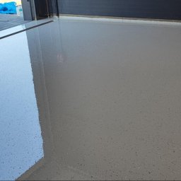 Piotr Cichoń - Błyszcząca, jasnoszara posadzka z drobnymi plamkami, świeżo wylana w garażu. Widoczne odbicie światła i fragment bramy garażowej.