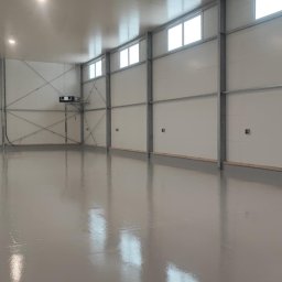 Piotr Cichoń - Aplikacja jasnoszarej, błyszczącej powłoki na posadzce w hali przemysłowej. Widoczny pracownik z wałkiem i puszką farby. Metalowa konstrukcja ścian i okna w tle.