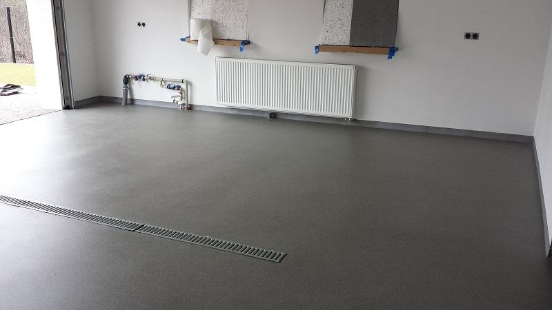 Szara, gładka podłoga z odpływem liniowym w nowym pomieszczeniu z grzejnikiem i elementami instalacji na ścianie, przygotowana do dalszych prac wykończeniowych.