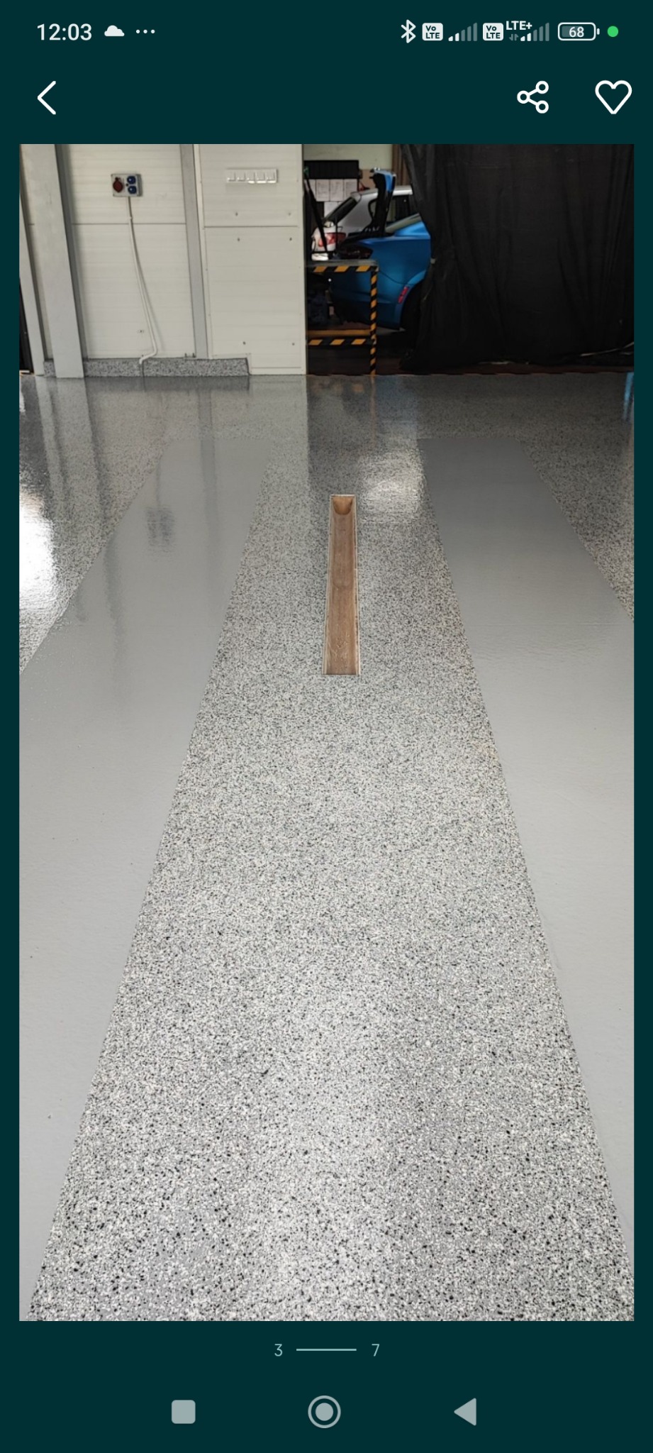 Szara, przemysłowa posadzka z żywicy z widocznym odpływem liniowym w garażu samochodowym, w tle częściowo widoczny niebieski samochód.