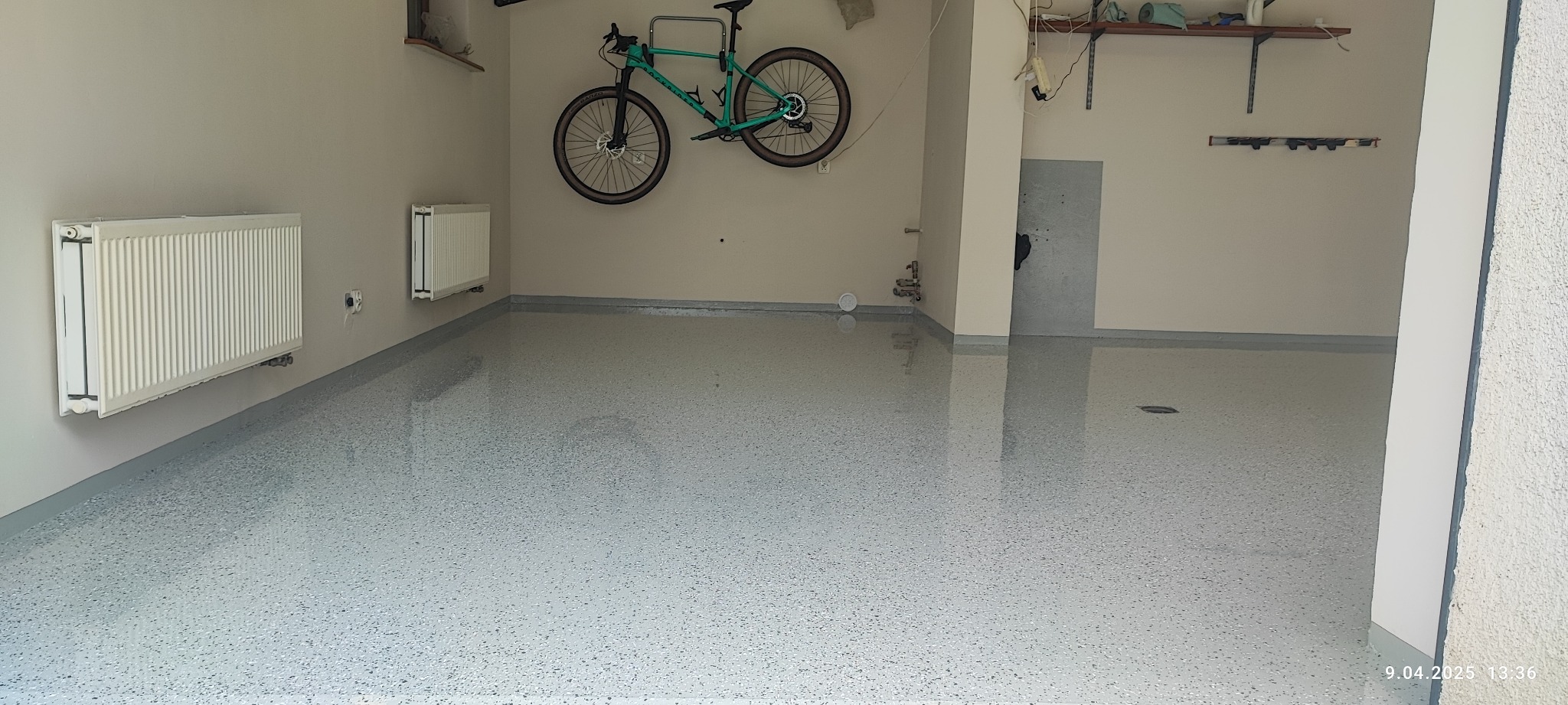 Jasnoszara, błyszcząca podłoga z żywicy z delikatnym wzorem, w garażu z rowerem wiszącym na ścianie i dwoma grzejnikami.