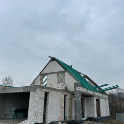 Dominik Błachut HOUSE-BUD - Surowy stan domu z betonu komórkowego, widoczne ściany, otwory okienne i drzwiowe, oraz elementy konstrukcji dachu z zielonymi łatami, na zewnątrz sterty gruzu i paleta z bloczkami, pochmurne niebo...