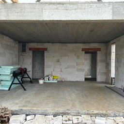 Dominik Błachut HOUSE-BUD - Surowy stan wnętrza domu w trakcie budowy: ściany z białych bloczków, betonowy strop, widoczne otwory drzwiowe, materiały budowlane i narzędzia na podłodze.