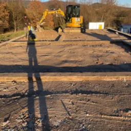 Przygotowanie terenu pod budowę: zagęszczarka wibracyjna w użyciu, minikoparka CAT w tle, widoczne zbrojenie i fundamenty, cień pracownika