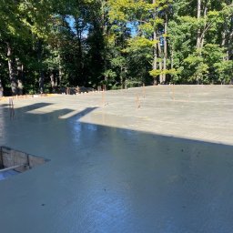 Dominik Błachut HOUSE-BUD - Wylana płyta fundamentowa z wystającymi prętami zbrojeniowymi, otoczona drewnianym szalunkiem, w tle drzewa.