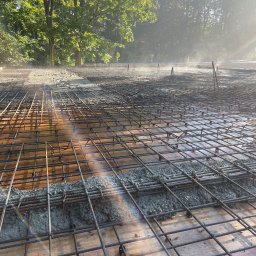 Dominik Błachut HOUSE-BUD - Zbrojenie fundamentu z częściowo wylanym betonem, widoczne stalowe pręty i drewniane szalunki, w tle zieleń drzew w porannym słońcu.