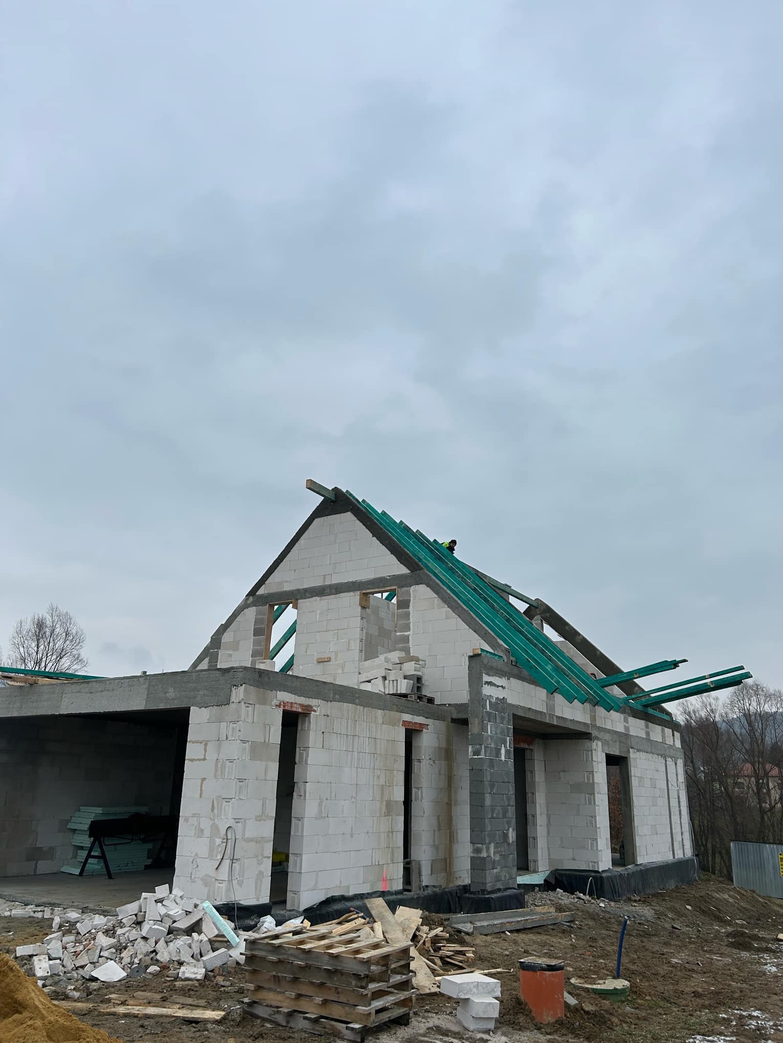 Surowy stan domu z betonu komórkowego, widoczne ściany, otwory okienne i drzwiowe, oraz elementy konstrukcji dachu z zielonymi łatami, na zewnątrz sterty gruzu i paleta z bloczkami, pochmurne niebo...