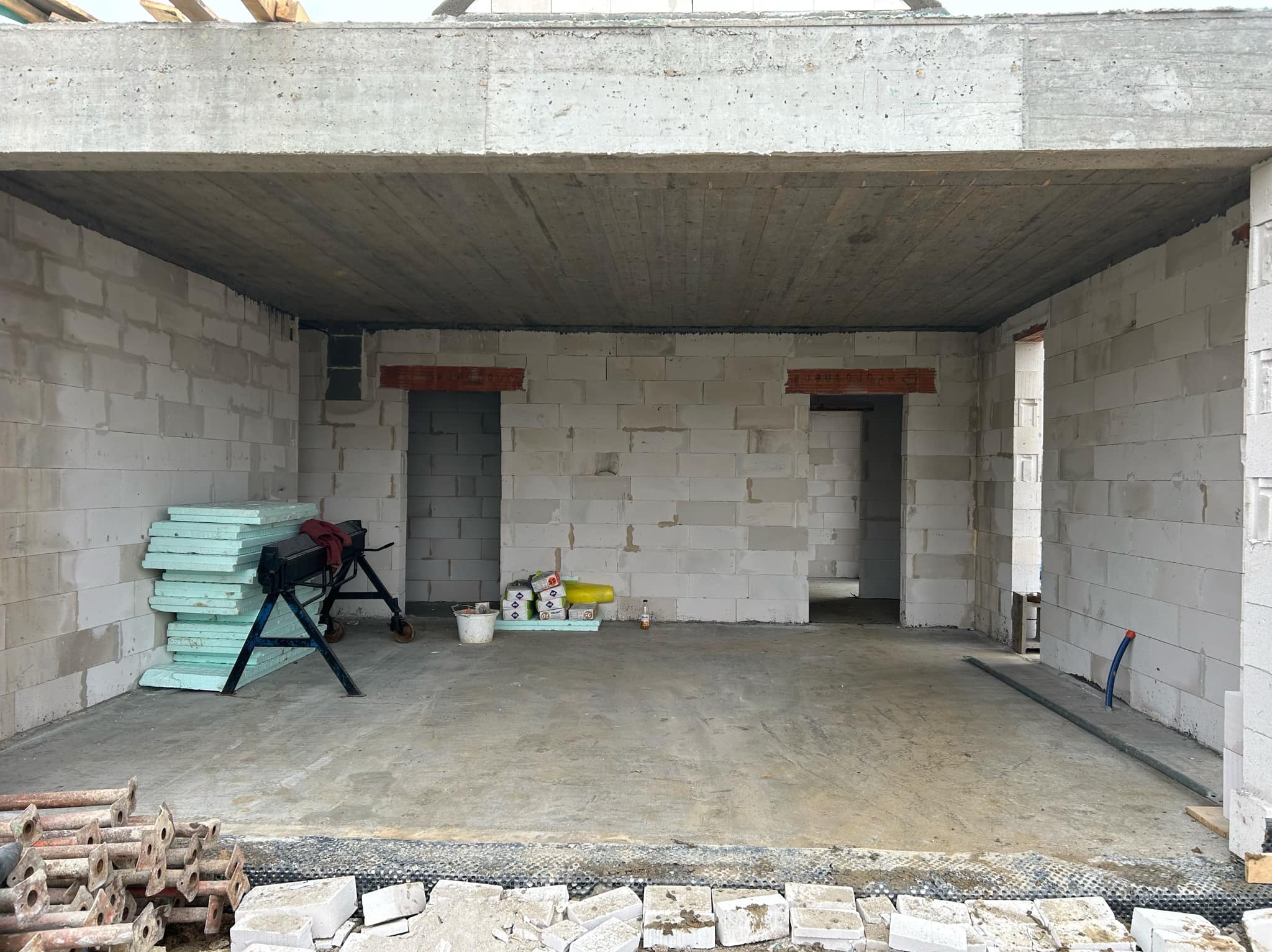 Surowy stan wnętrza domu w trakcie budowy: ściany z białych bloczków, betonowy strop, widoczne otwory drzwiowe, materiały budowlane i narzędzia na podłodze.