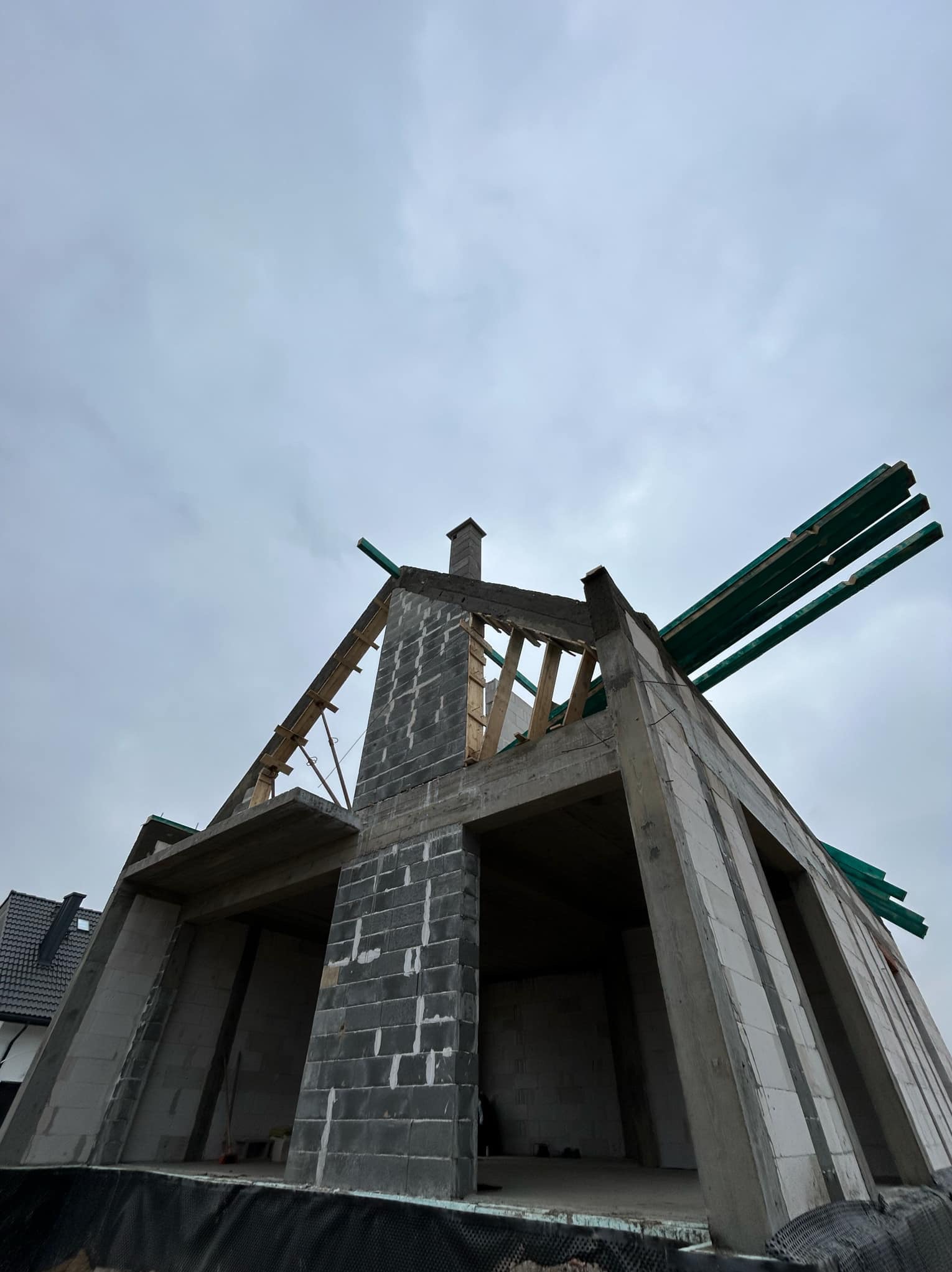 Surowy stan domu murowanego w trakcie budowy, widok z dołu na ściany z betonu komórkowego, drewniane elementy konstrukcji dachu i komin z cegły, pochmurne niebo w tle.