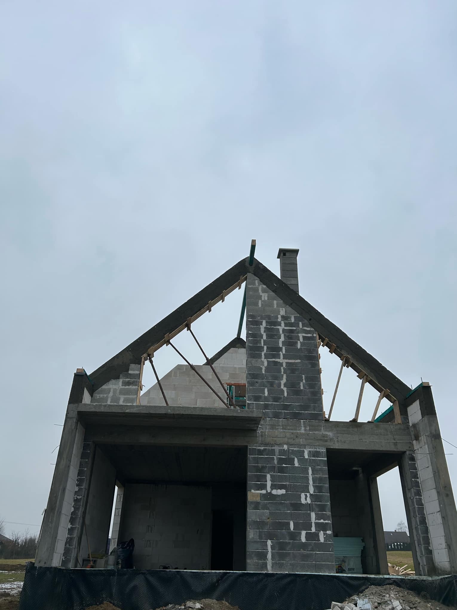 Surowy stan domu murowanego z kominem, widok z dołu na tle pochmurnego nieba, brakujące elementy dachu i okien.