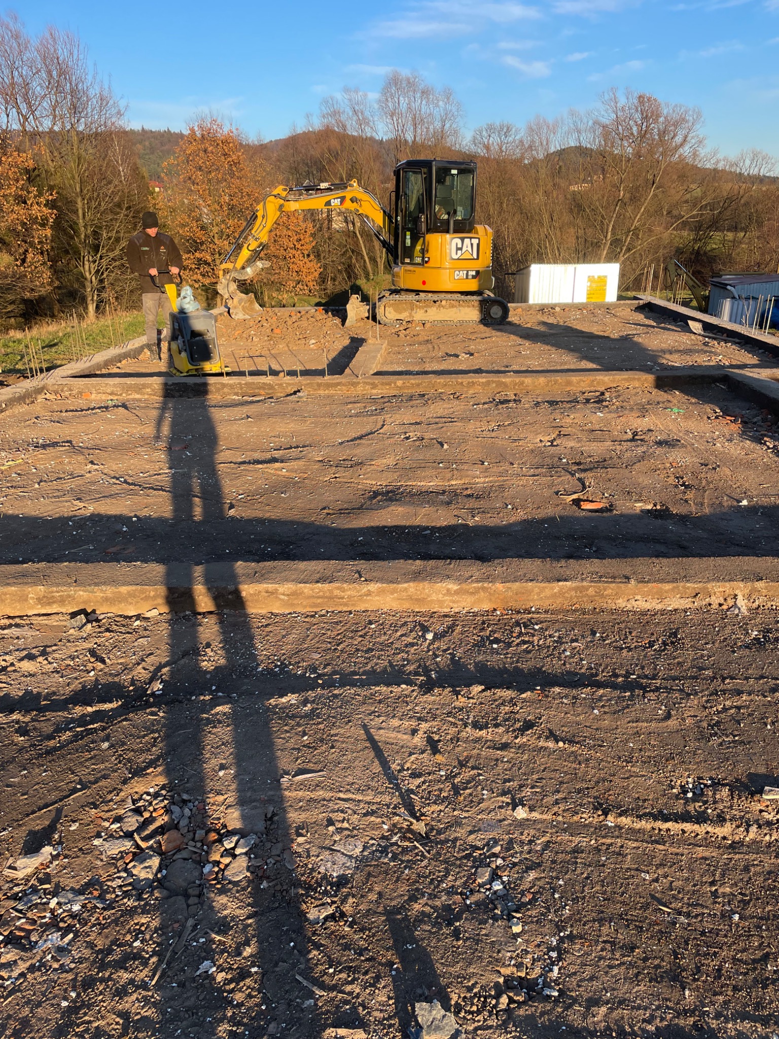 Przygotowanie terenu pod budowę: zagęszczarka wibracyjna w użyciu, minikoparka CAT w tle, widoczne zbrojenie i fundamenty, cień pracownika