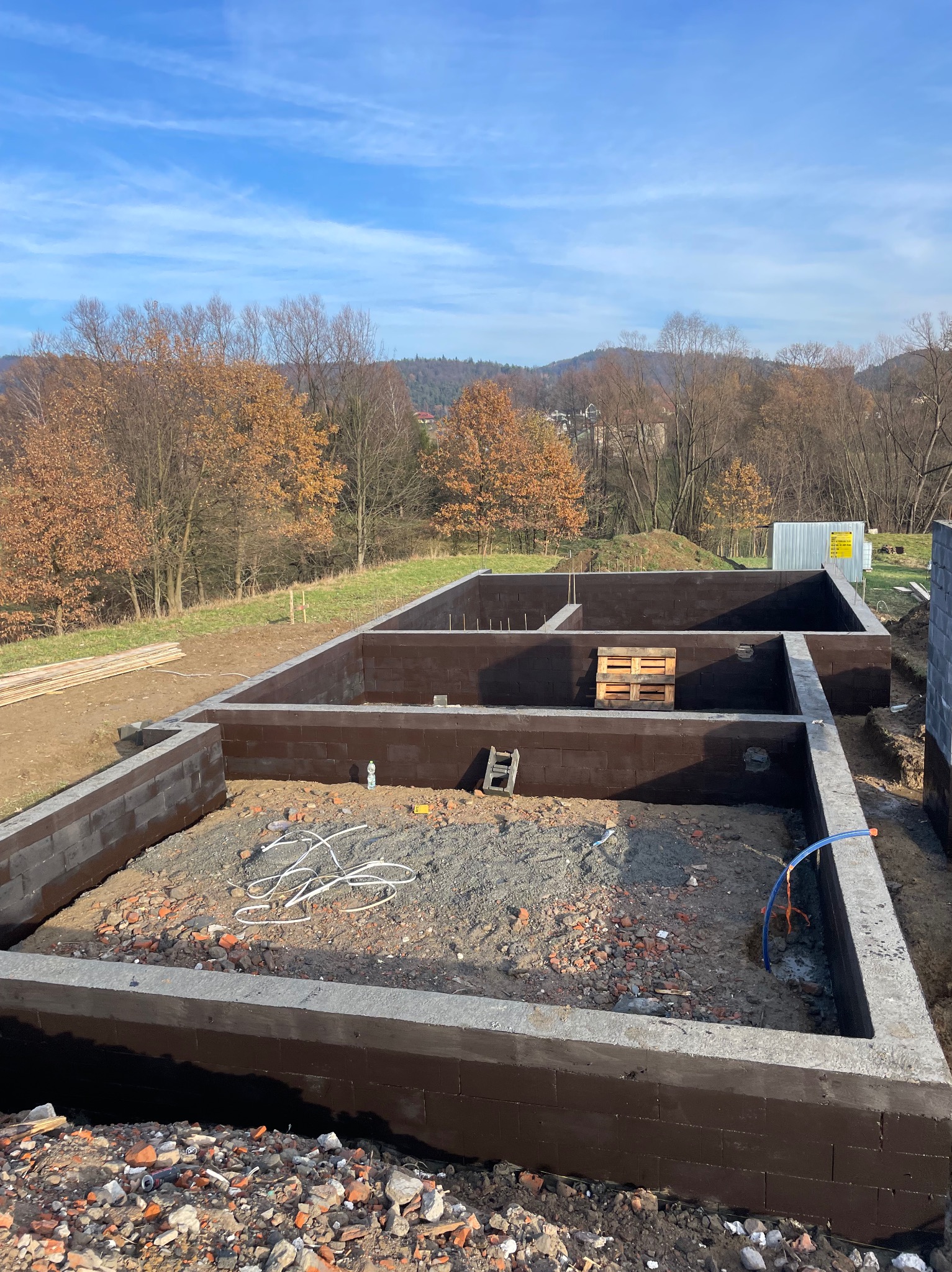 Wykopane i częściowo wylane fundamenty pod dom z widocznymi bloczkami fundamentowymi, izolacją przeciwwilgociową, gruzem, przewodami elektrycznymi, paletą drewnianą, drabinką oraz otoczeniem...