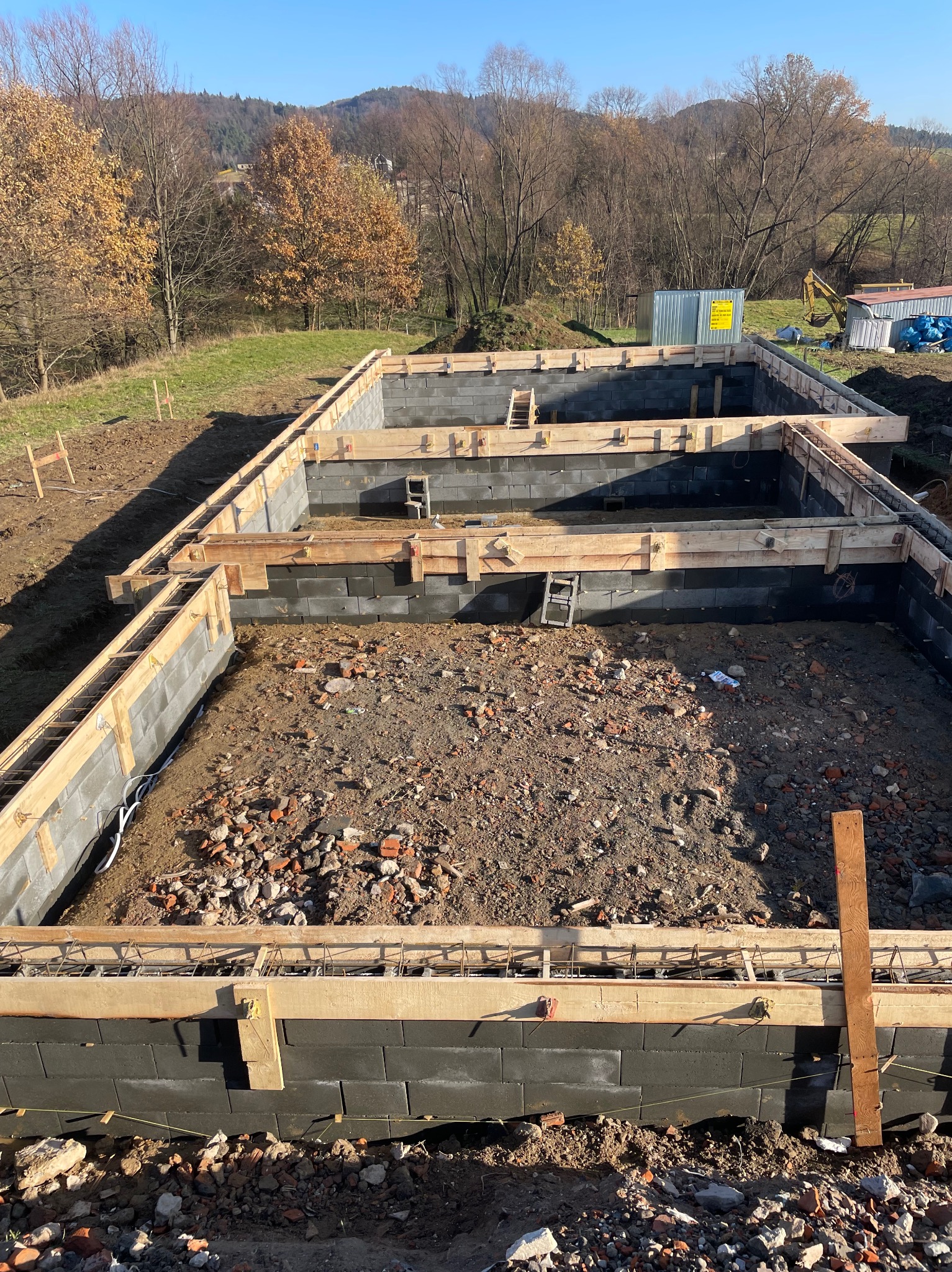 Fundamenty domu w trakcie budowy, widoczne bloczki betonowe, drewniane szalunki i zbrojenie, w tle krajobraz zalesionych wzgórz.