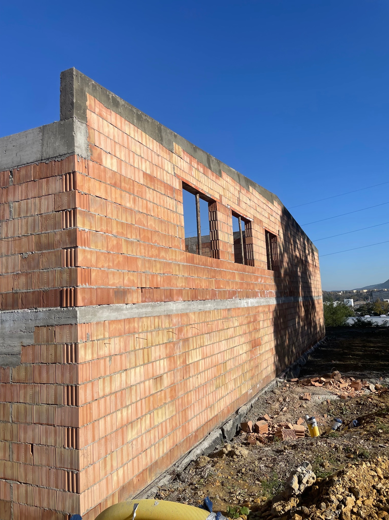 Wznoszenie ścian domu z czerwonej ceramiki poryzowanej, widoczne otwory okienne i fragmenty betonowego wieńca, na zewnątrz budynku leżą gruz i żółty wąż wentylacyjny.