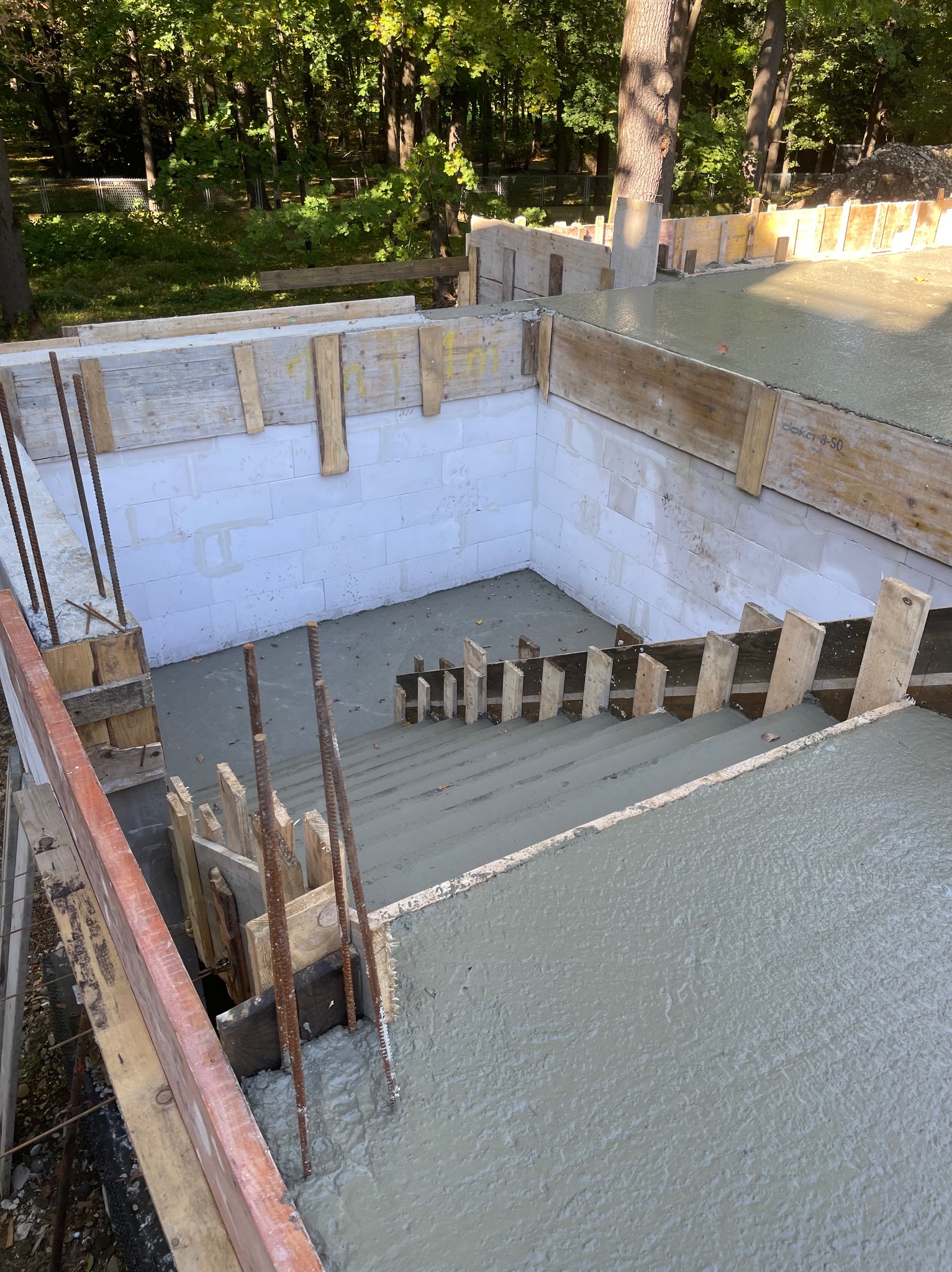 Świeżo wylany beton fundamentowy w szalunkach z drewnianych desek i zbrojeniem stalowym, widok z góry na teren budowy z drzewami w tle.
