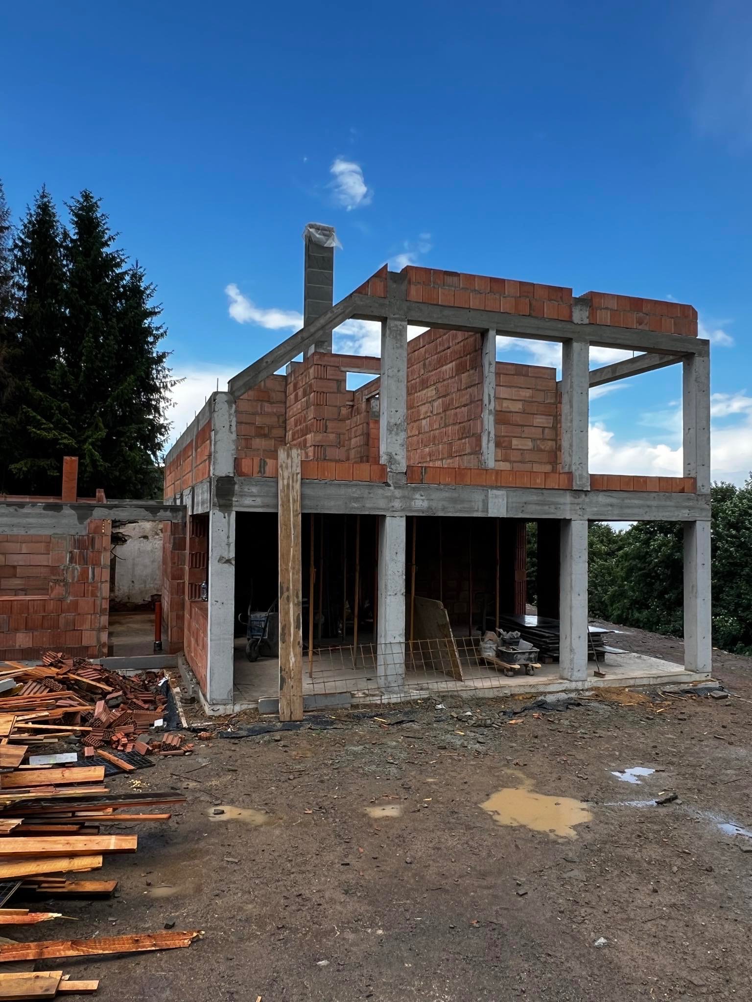 Surowy stan dwukondygnacyjnego domu z czerwonej cegły i betonu w trakcie budowy, widoczne fragmenty szalunków i sterty materiałów budowlanych na placu budowy pod błękitnym niebem.