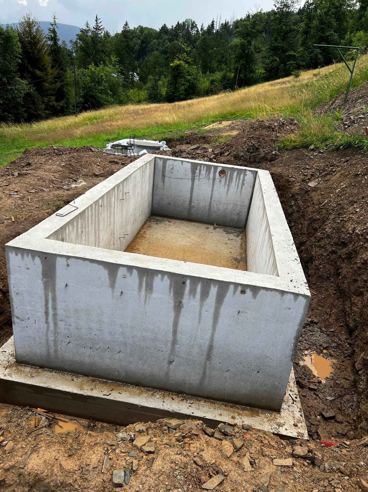 Betonowa konstrukcja fundamentu w wykopie na tle zielonego wzgórza z drzewami, widoczne ślady wilgoci na ścianach betonu.