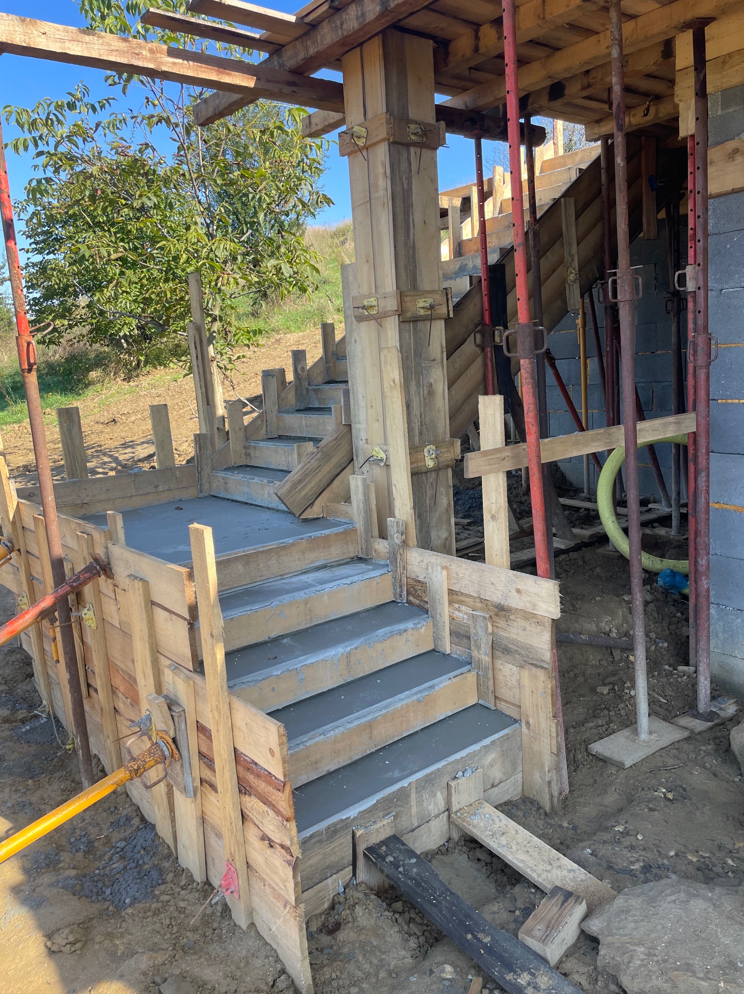 Betonowe schody w drewnianym szalunku podczas budowy, widoczne podpory stalowe, fragment ściany z bloczków i drzewo w tle.