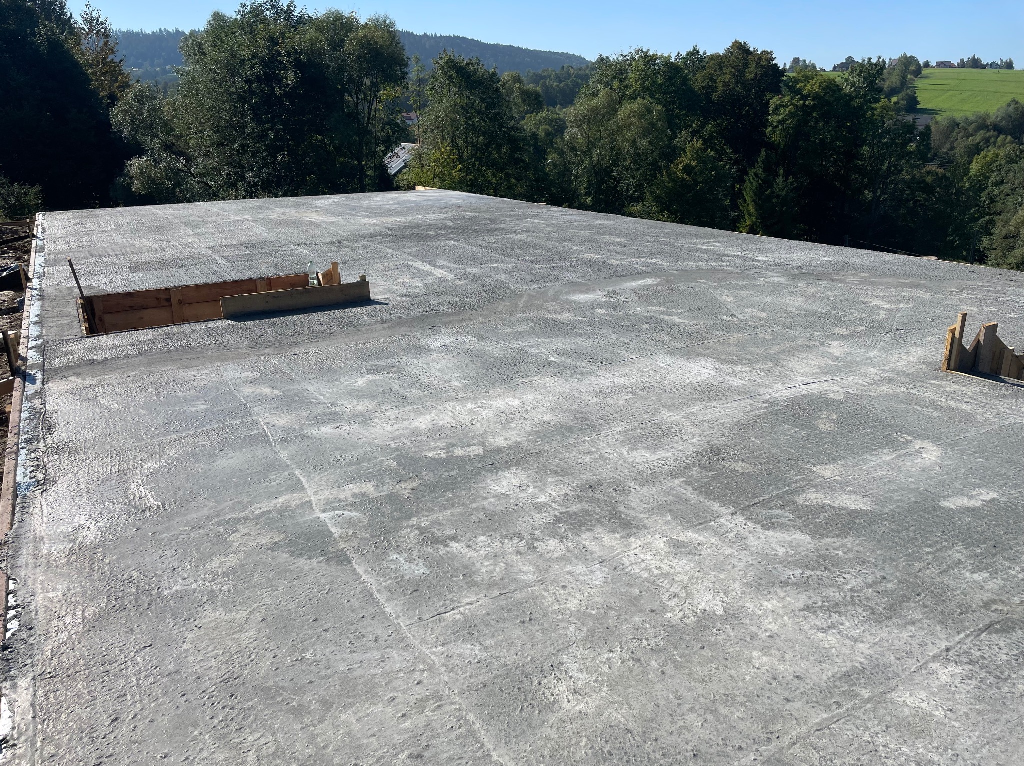 Zalany, świeży fundament płyty betonowej z drewnianym szalunkiem w Krzeszowie, z widokiem na zalesione wzgórza w tle.