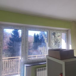 KRAFTVIT SPÓŁKA Z OGRANICZONĄ ODPOWIEDZIALNOŚCIĄ - Pokój w trakcie remontu: zielone ściany z widocznymi śladami po pracach, okno balkonowe z widokiem na drzewa, zakurzony grzejnik, karton na szafce i wałek do malowania oparty o ścianę.