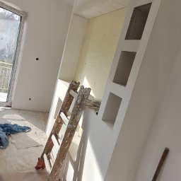 KRAFTVIT SPÓŁKA Z OGRANICZONĄ ODPOWIEDZIALNOŚCIĄ - Ściana z wnękami w trakcie wykańczania, widoczne ślady po gładzi, drabina oparta o ścianę, podłoga częściowo zakryta folią malarską, widoczne narzędzia malarskie.