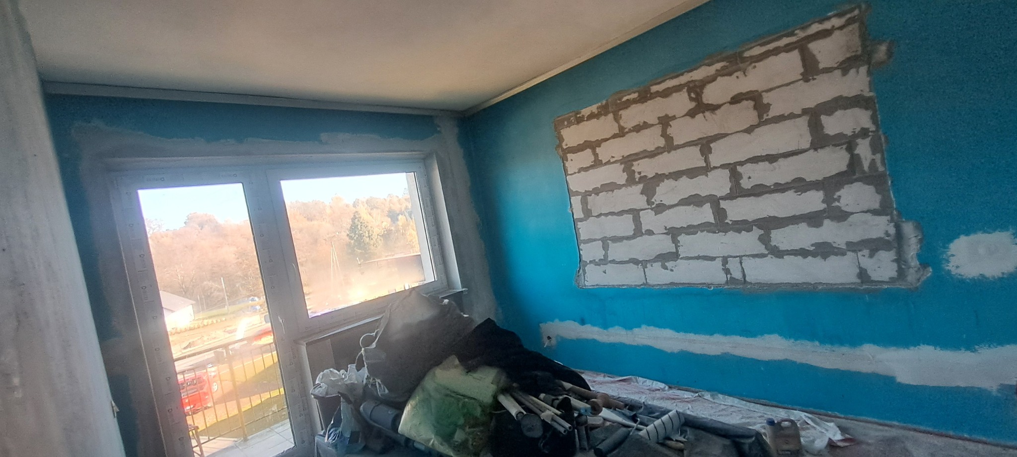 Niebieska ściana z częściowo zamurowanym oknem z białej cegły, widoczne ślady po gładzi i malowaniu, w rogu sterta narzędzi i materiałów budowlanych.