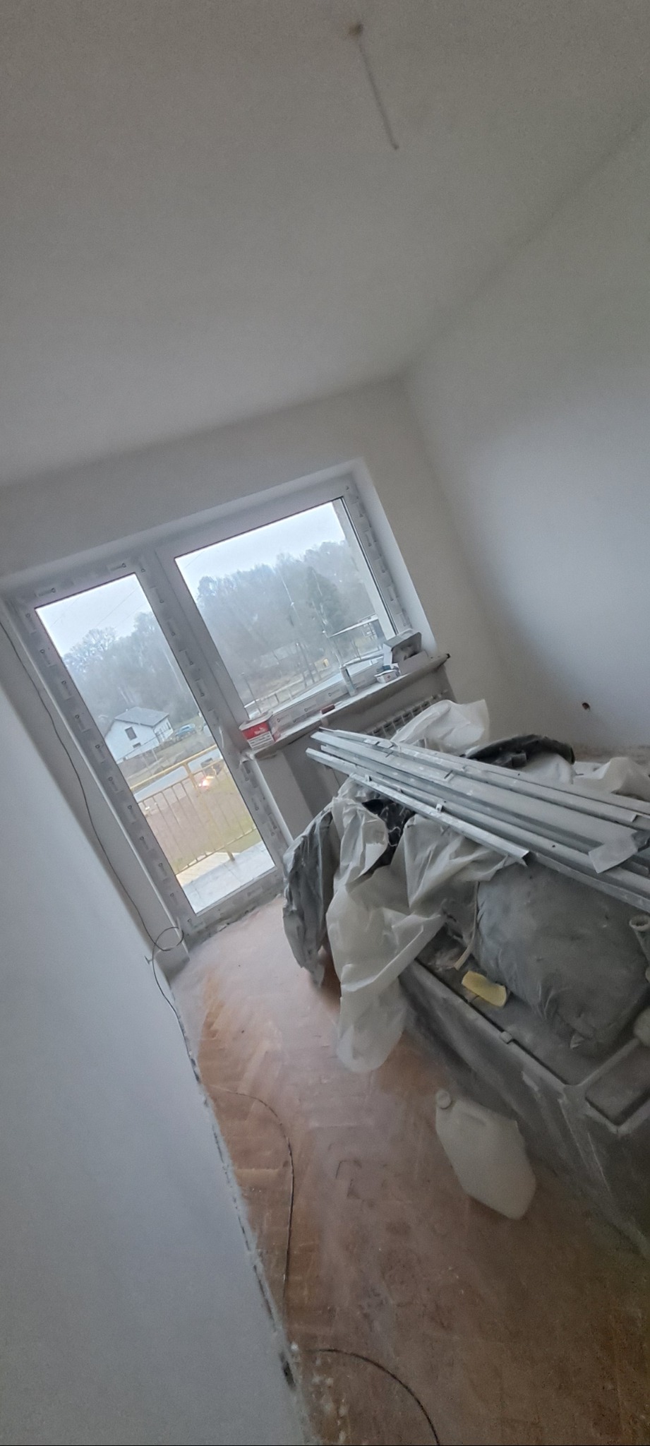 Pokój w trakcie remontu: świeżo pomalowane ściany na biało, okno z widokiem na zewnątrz, podłoga częściowo zakryta, profile aluminiowe i pojemnik z farbą widoczne.