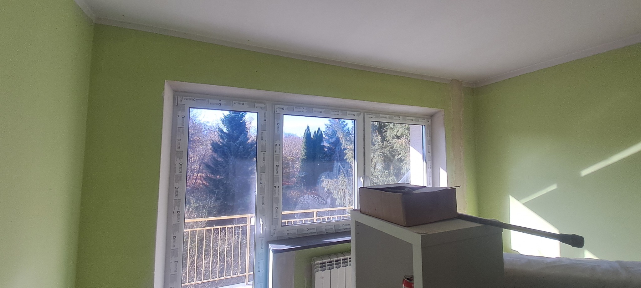 Pokój w trakcie remontu: zielone ściany z widocznymi śladami po pracach, okno balkonowe z widokiem na drzewa, zakurzony grzejnik, karton na szafce i wałek do malowania oparty o ścianę.
