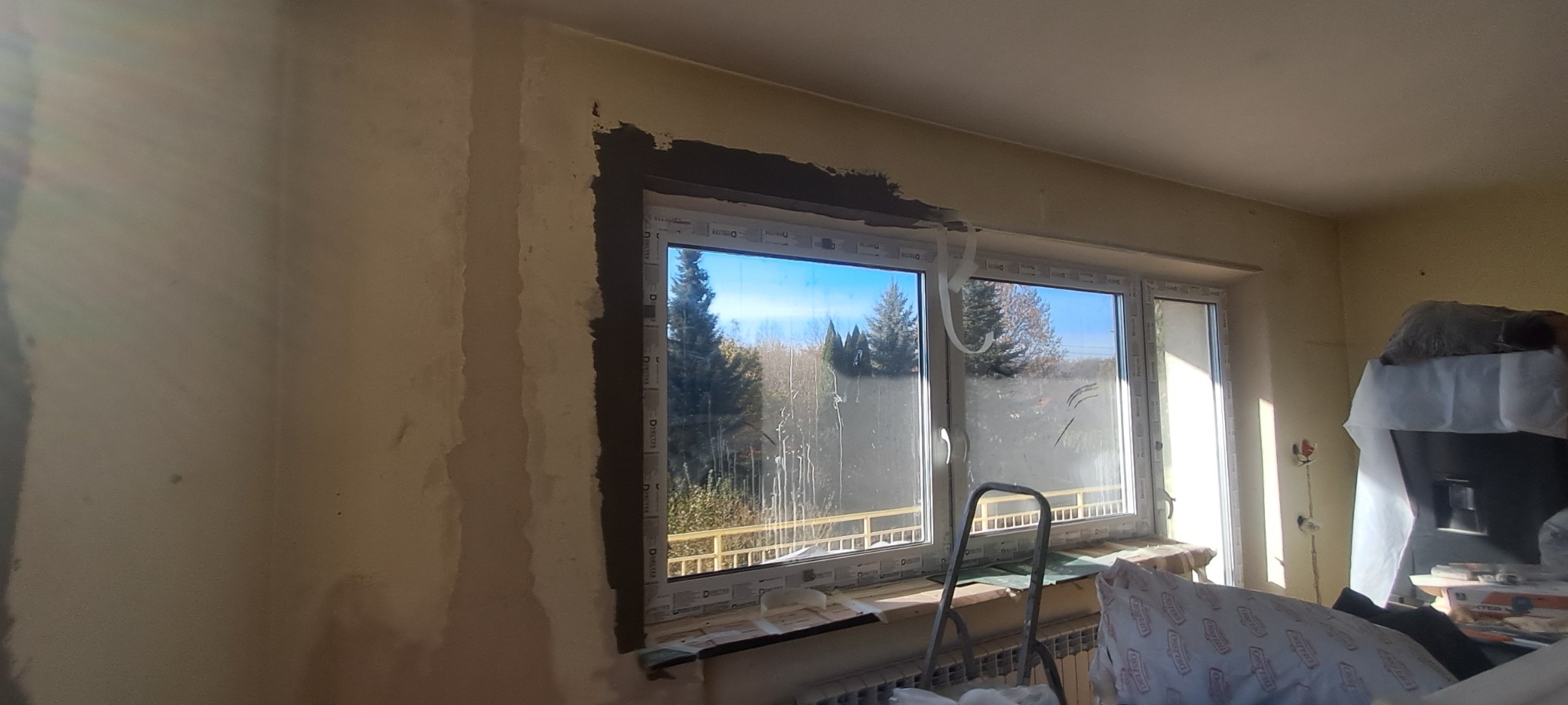 Przygotowanie ściany do gładzi wokół okna PCV, widoczne surowe wykończenie i drabina w pokoju