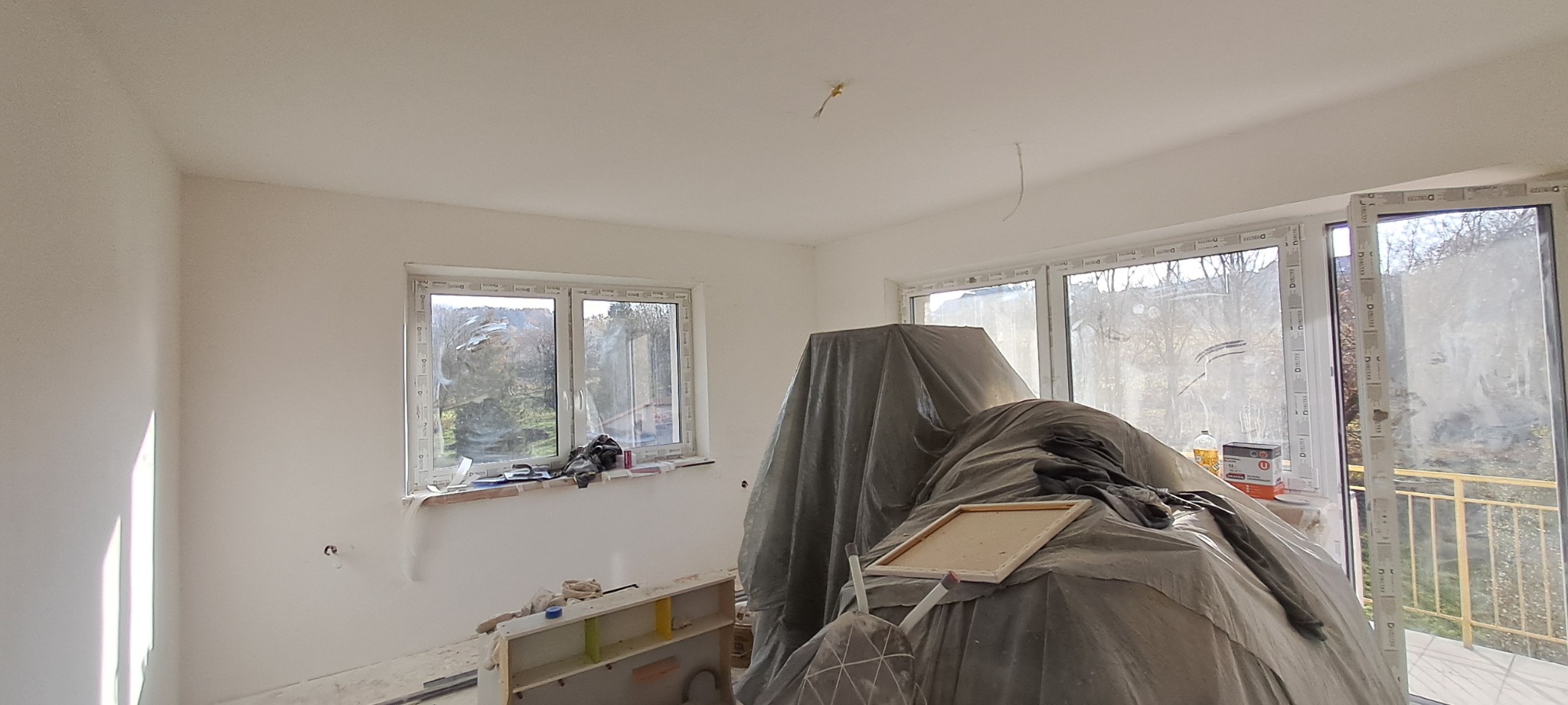 Pokój w trakcie remontu: świeżo położone gładzie na ścianach, okna zabezpieczone folią, sprzęt budowlany przykryty plandeką, widoczne niedociągnięcia i resztki materiałów.