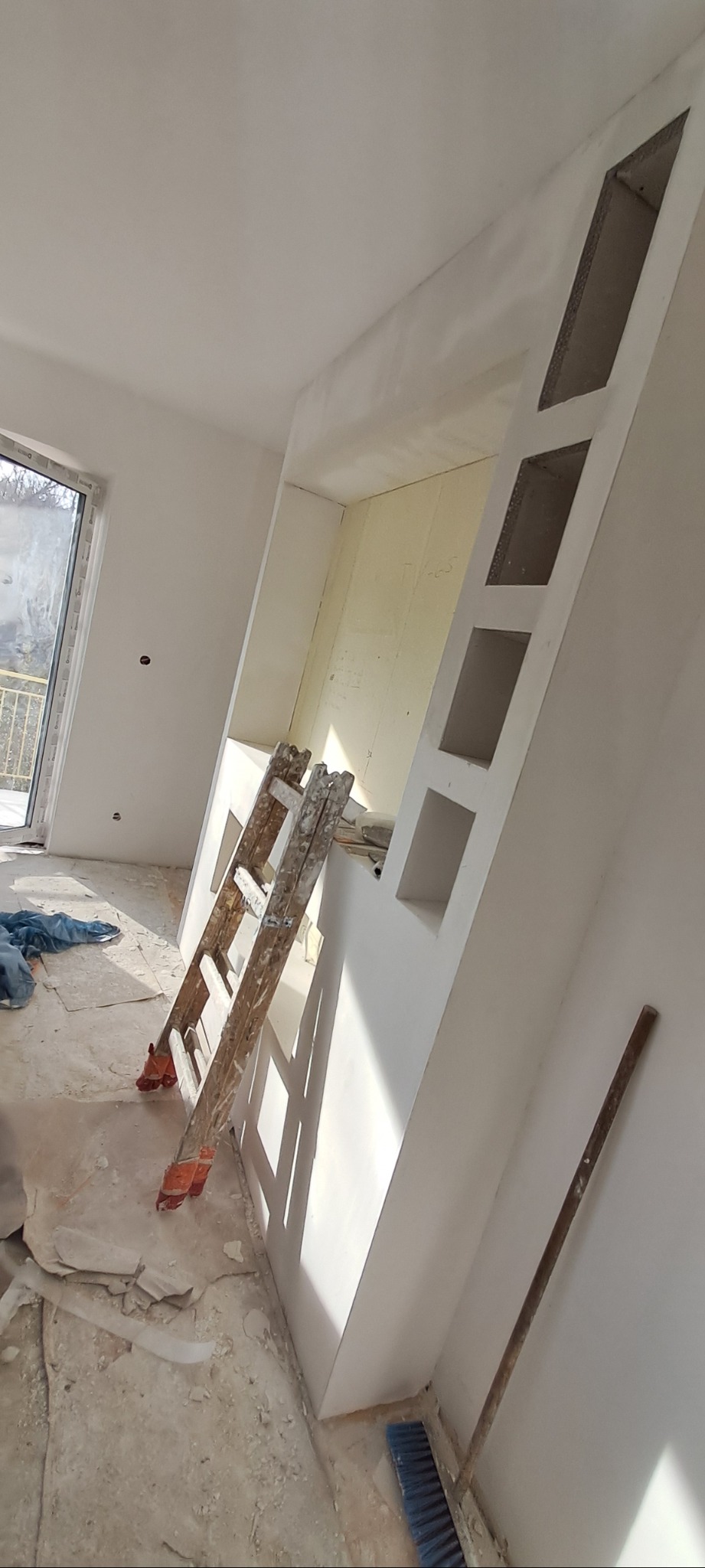 Ściana z wnękami w trakcie wykańczania, widoczne ślady po gładzi, drabina oparta o ścianę, podłoga częściowo zakryta folią malarską, widoczne narzędzia malarskie.