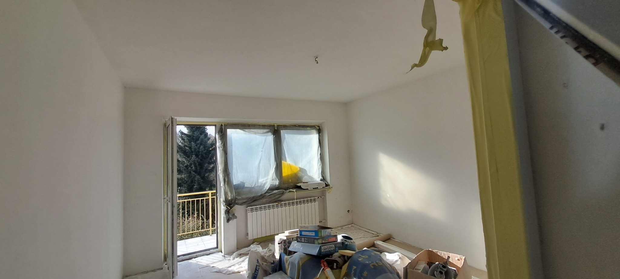 Pokój w trakcie przygotowań do malowania, z zabezpieczonymi oknami folią i taśmą, świeżo zagruntowanymi ścianami oraz widocznymi narzędziami i materiałami malarskimi.