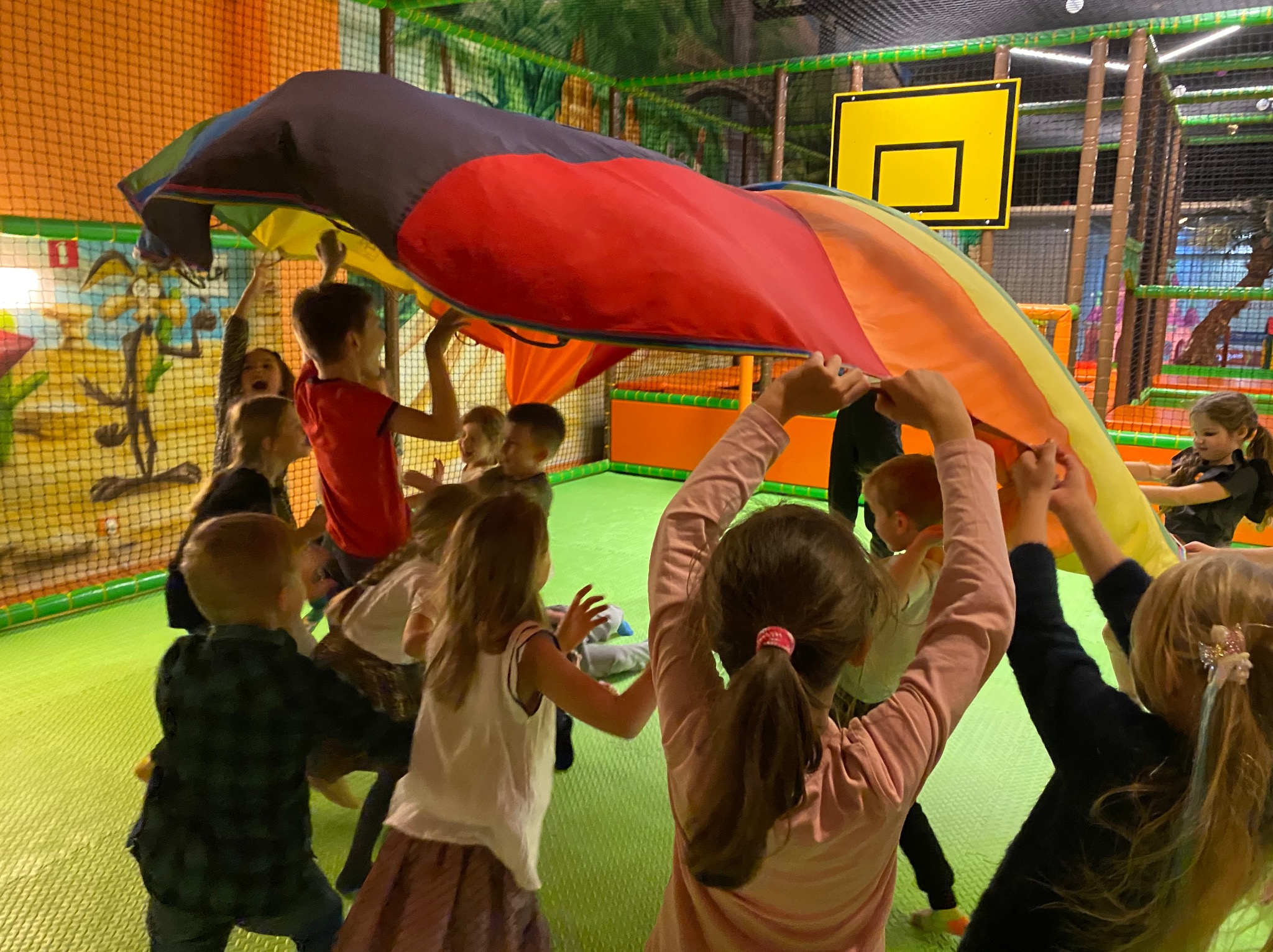 Grupa dzieci w sali zabaw trzymająca kolorową chustę animacyjną nad głowami, w tle widoczna konstrukcja placu zabaw z motywem dżungli i koszem do koszykówki.