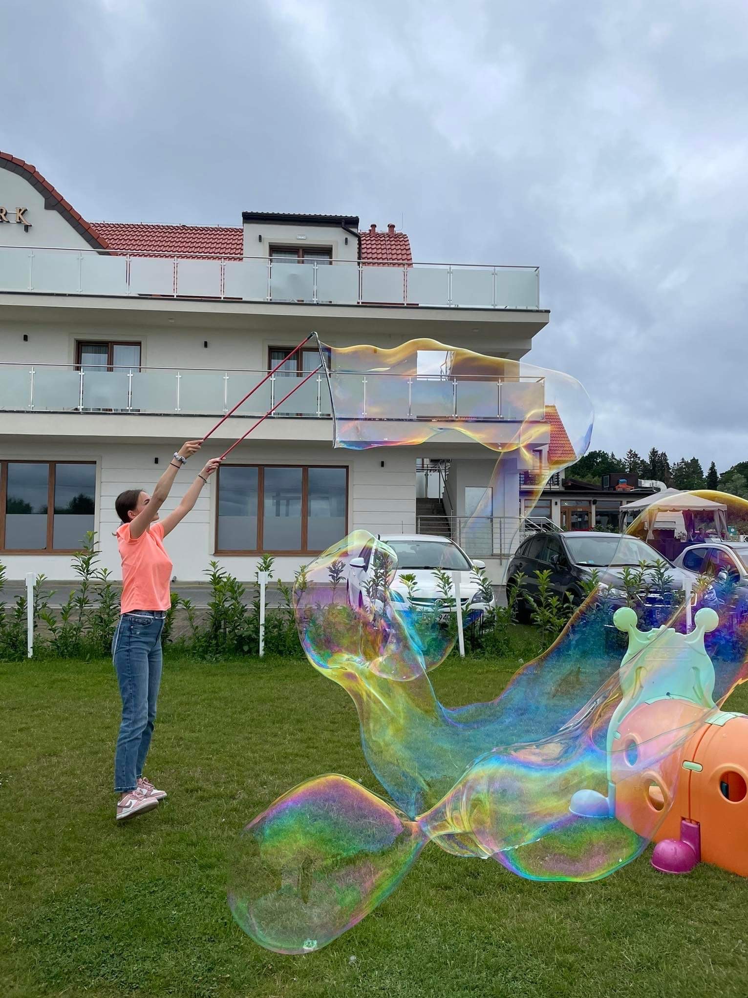 Dziewczyna w różowej koszulce puszcza ogromne, tęczowe bańki mydlane za pomocą specjalnego sznurka na trawniku przed białym budynkiem z czerwonym dachem.