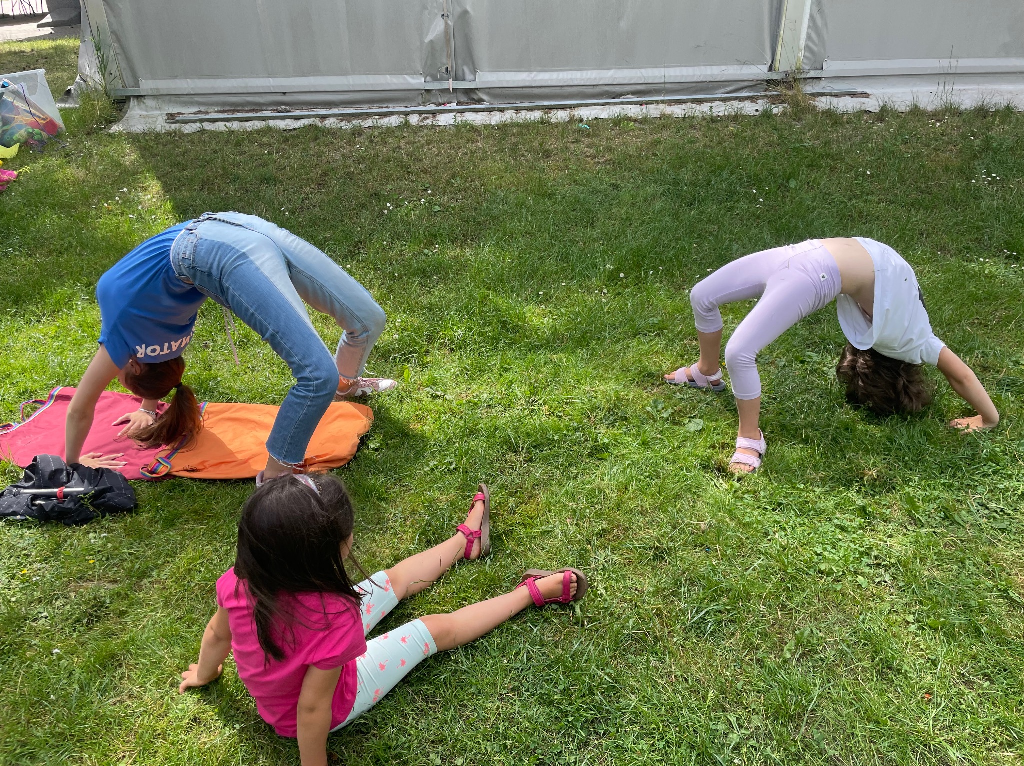 Dwie dziewczynki w pozycji mostka na trawie, trzecia siedzi obok, w tle namiot