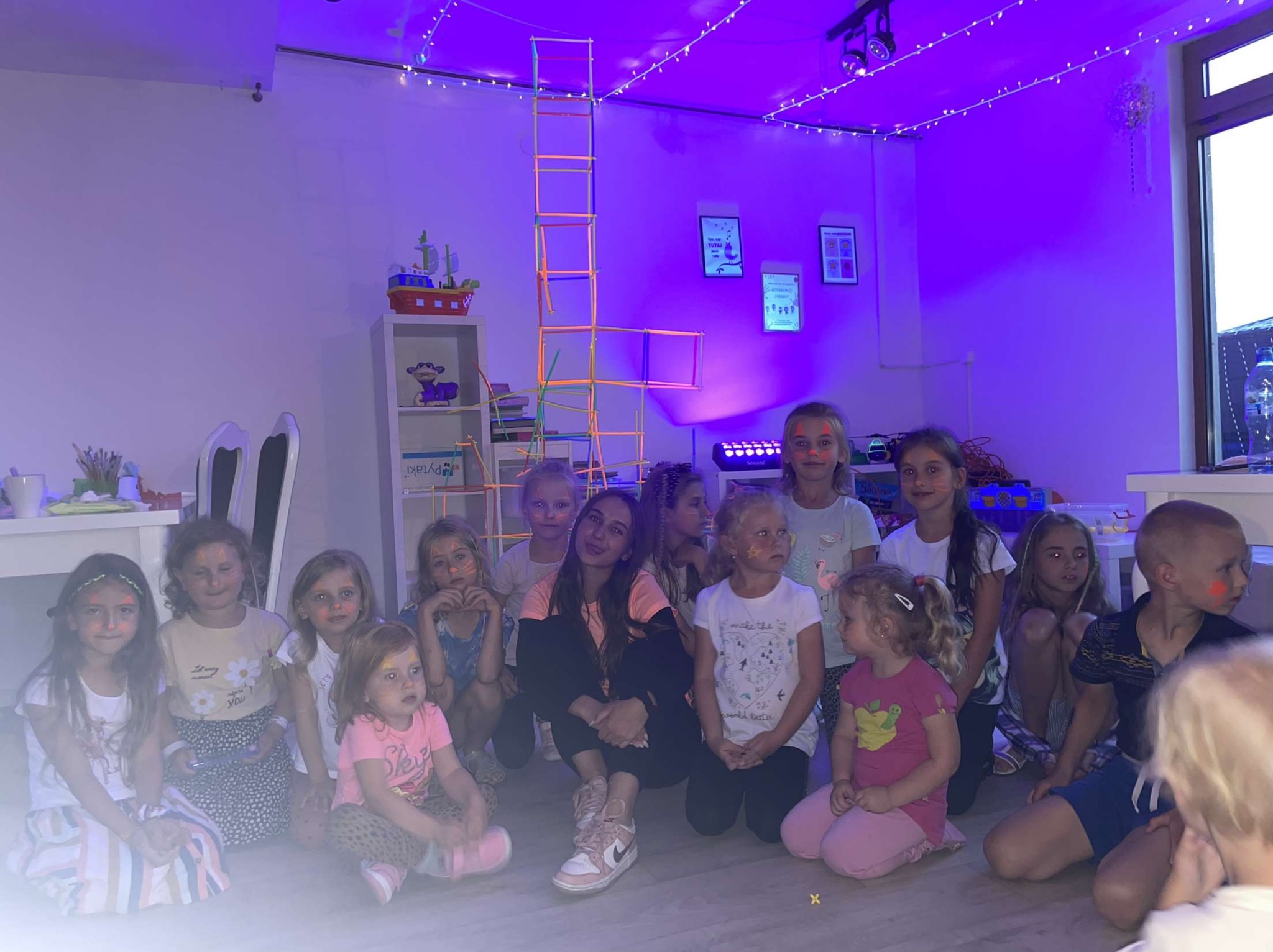 Grupa dzieci z pomalowanymi twarzami i animatorka siedząca na podłodze w pokoju oświetlonym fioletowym światłem, w tle konstrukcja z rurek i półka z zabawkami.