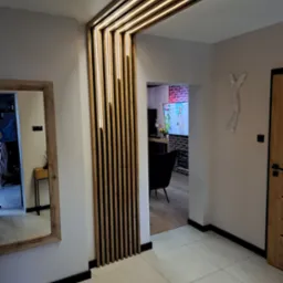 Nowoczesne oświetlenie LED w drewnianej konstrukcji oddzielającej przestrzeń w przedpokoju. Lustro z drewnianą ramą i płytki podłogowe w neutralnej kolorystyce.