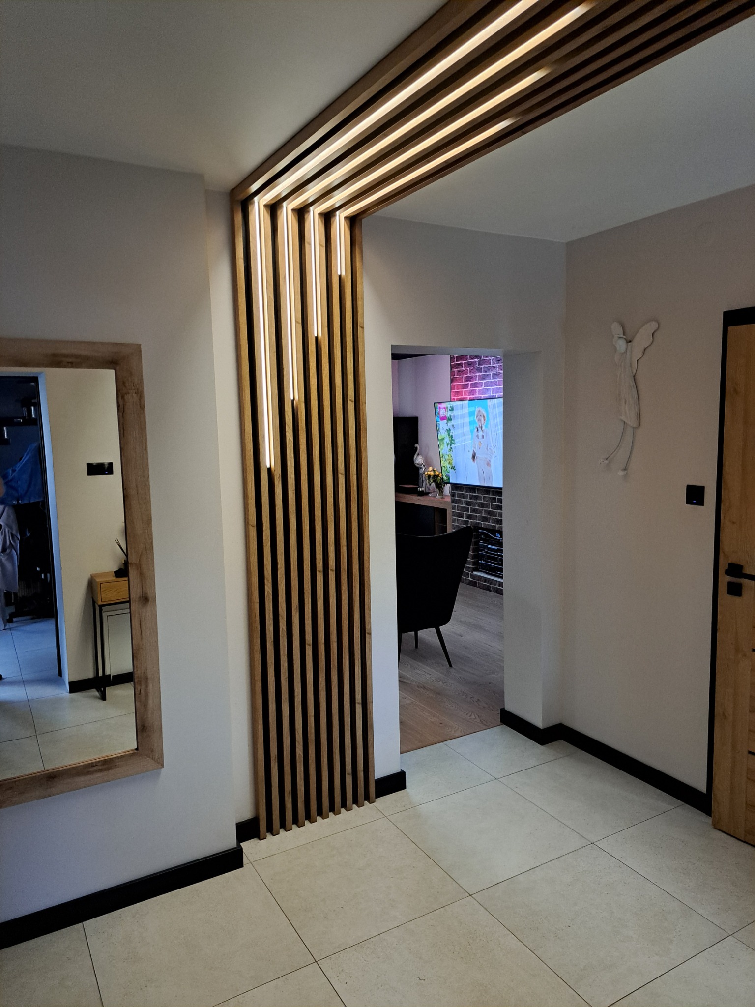 Nowoczesne oświetlenie LED w drewnianej konstrukcji oddzielającej przestrzeń w przedpokoju. Lustro z drewnianą ramą i płytki podłogowe w neutralnej kolorystyce.