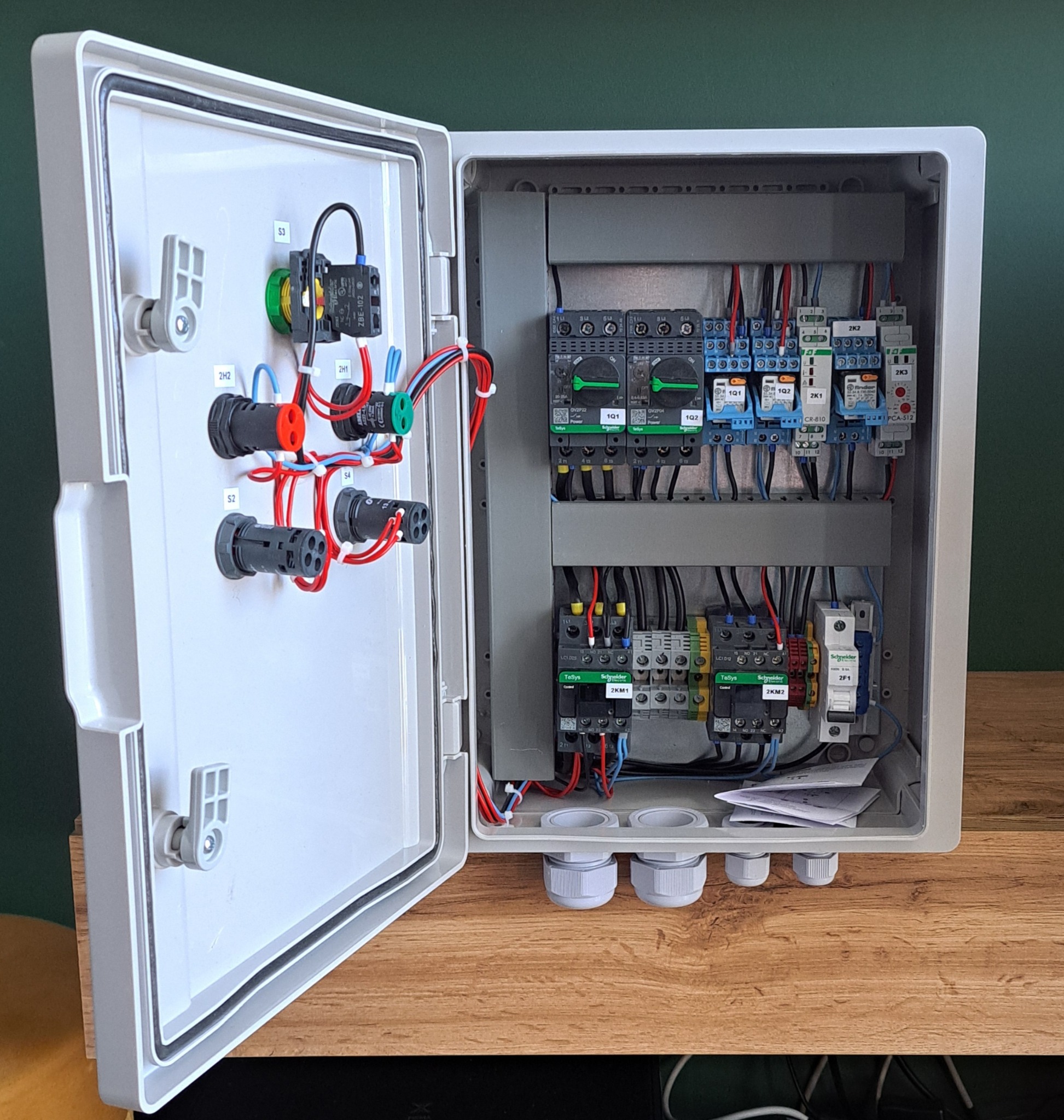 Otwarta skrzynka elektryczna z widocznymi bezpiecznikami, przełącznikami i okablowaniem, zamontowana na drewnianej powierzchni. Widoczne elementy sterownicze na panelu.