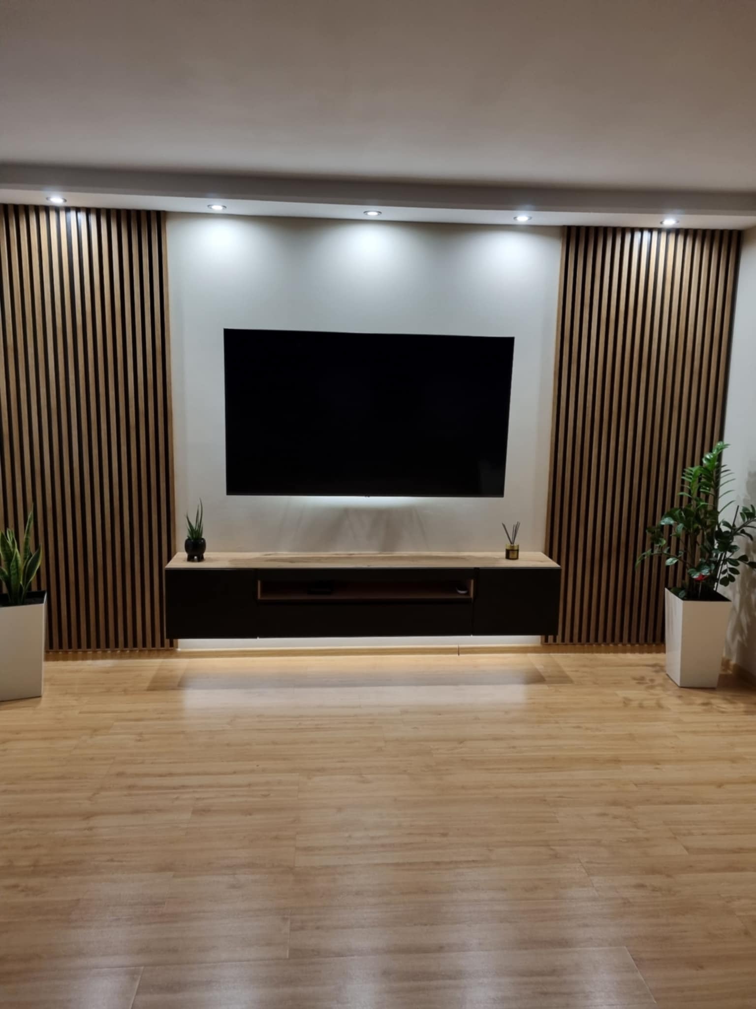 Minimalistyczny salon z drewnianymi lamelami na ścianie, podświetlaną szafką RTV i dużym telewizorem, dopełniony roślinami w białych donicach.