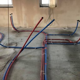 ADAM RADASZKIEWICZ Hydro-Instal - Instalacja ogrzewania podłogowego w trakcie montażu: rury PEX czerwone i niebieskie ułożone na surowym betonie, przymocowane plastikowymi uchwytami, widoczne otwory w podłodze na pionowe rury...