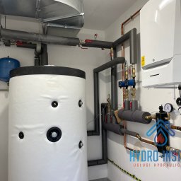 ADAM RADASZKIEWICZ Hydro-Instal - Biały zasobnik ciepłej wody użytkowej z izolacją, obok wiszący kocioł gazowy De Dietrich, rury miedziane i izolowane, dwa niebieskie zbiorniki wyrównawcze, widoczne logo firmy hydraulicznej...