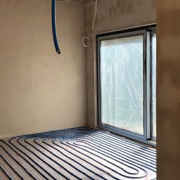 ADAM RADASZKIEWICZ Hydro-Instal - Niebieskie rury ogrzewania podłogowego ułożone na siatce w surowym pomieszczeniu z oknem zabezpieczonym folią, wisząca rura doprowadzająca.